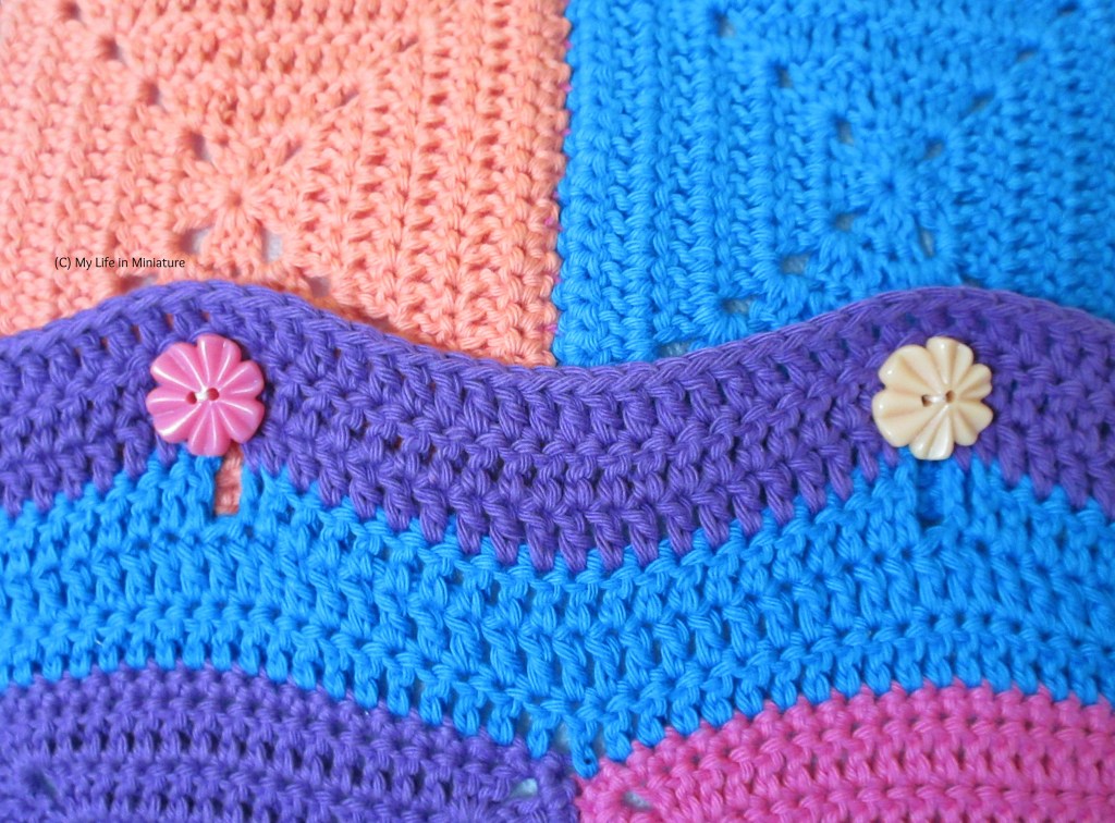 Close-up of two of the buttons on the cushion cover. The left button is dark pink, the right button is a pale peach. The crocheted overlap has buttonholes in it, and is striped blue and purple. 