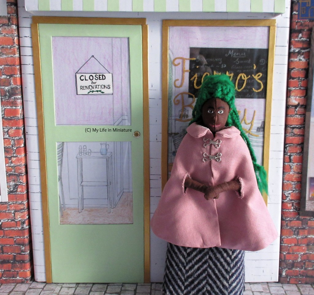 Hazel stands in front of the window outside Fierro's Bakery. She smiles at the camera, rubbing her hands together in front of her. The door is clearly visible, with two windows cut into it. In the bottom window is the back of a chair and a water cart. In the top window is a sign that says 'Closed for Renovations'. 