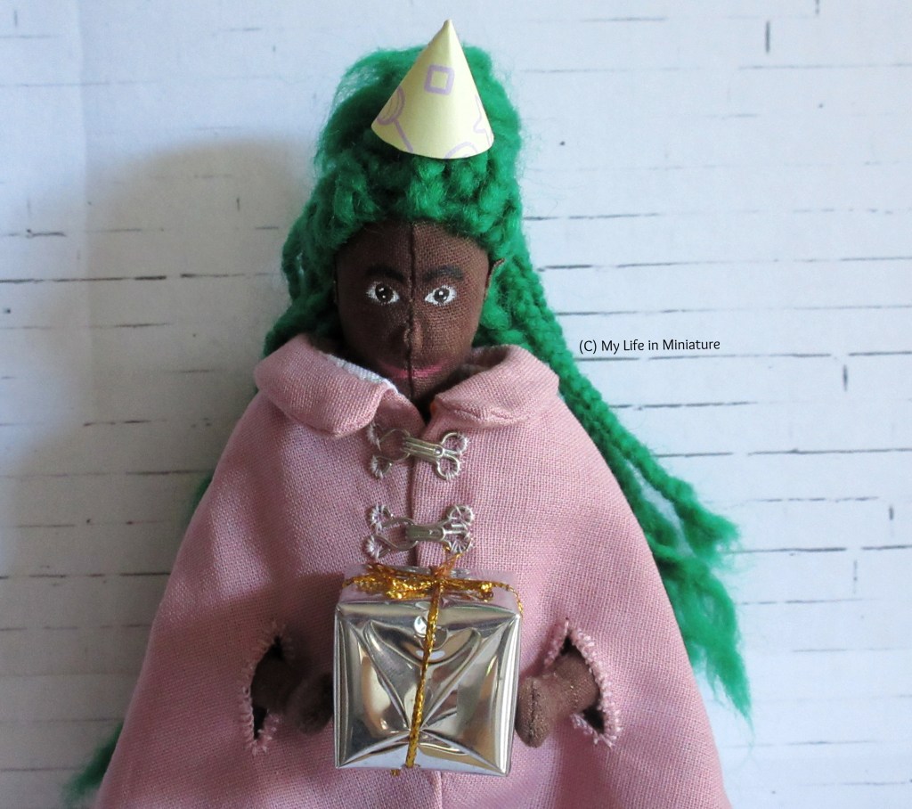 Hazel stands in front of a white brick background, holding a silver-wrapped present and wearing a conical party hat. She also wears a pale pink cape, and has her green braids down her back. She smiles down at the gift. 