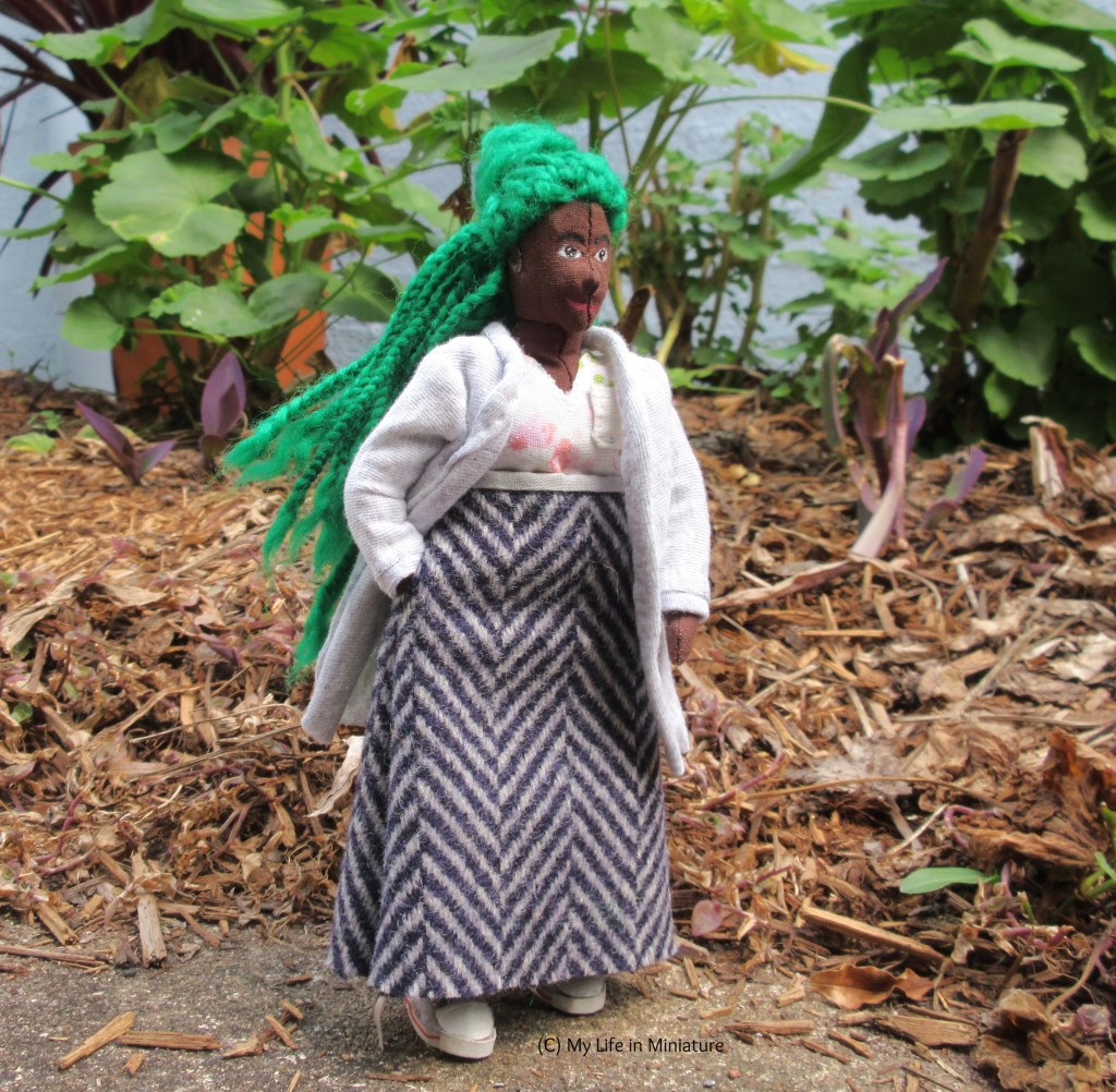 Hazel stands outside, wearing a grey jersey cardigan, a grey-and-navy wool skirt, and a pink floral V-necked top. One hand is in her skirt pocket, while the other hangs at her side. She looks to the right, and her braids lift slightly in the breeze. 