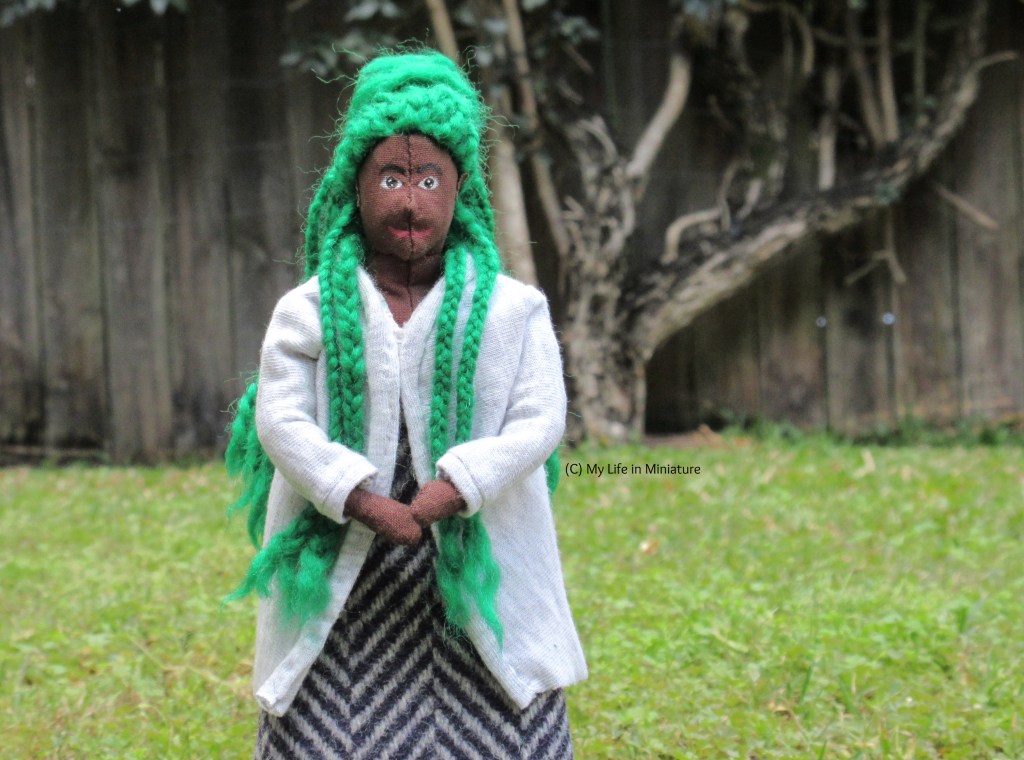 Hazel stands on grass, wearing a grey jersey cardigan and a grey-and-navy wool skirt. She has her hands clasped in front of her, and has two braids over the front of each shoulder. She smiles at the camera.
