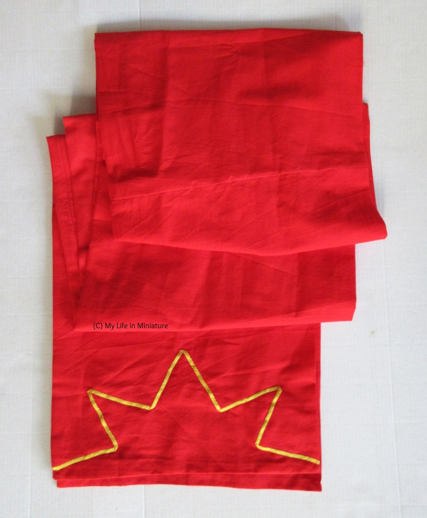 A long red scarf, folded to fit into frame, sits on a white background. A length of yellow ribbon is sewn onto each end in a half-star shape. 