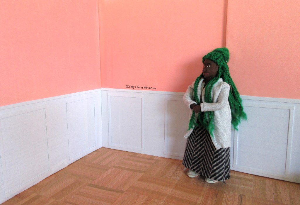 Hazel stands in a different area of the interior of Fierro's Bakery. She's dusting off her hands, wearing a wool skirt and grey cardigan. The top two-thirds of the walls are pale pink, and the bottom third is white with raised panelling. The floor is parquet wood.