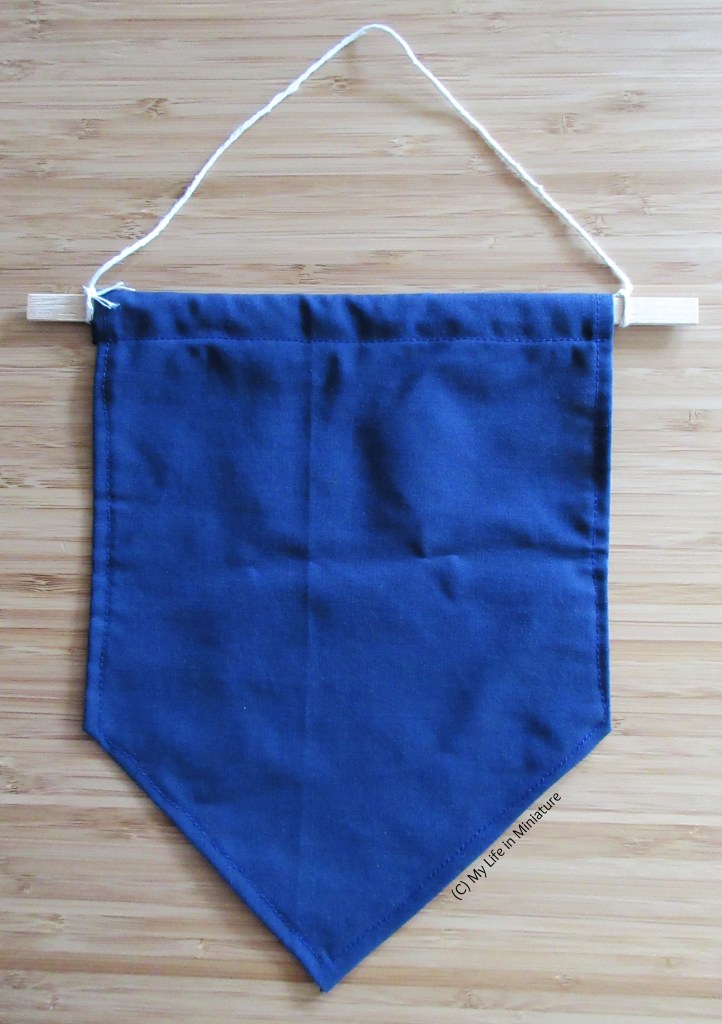 A small navy blue fabric banner is laid flat on a wood background, with the bar and string at the top. 