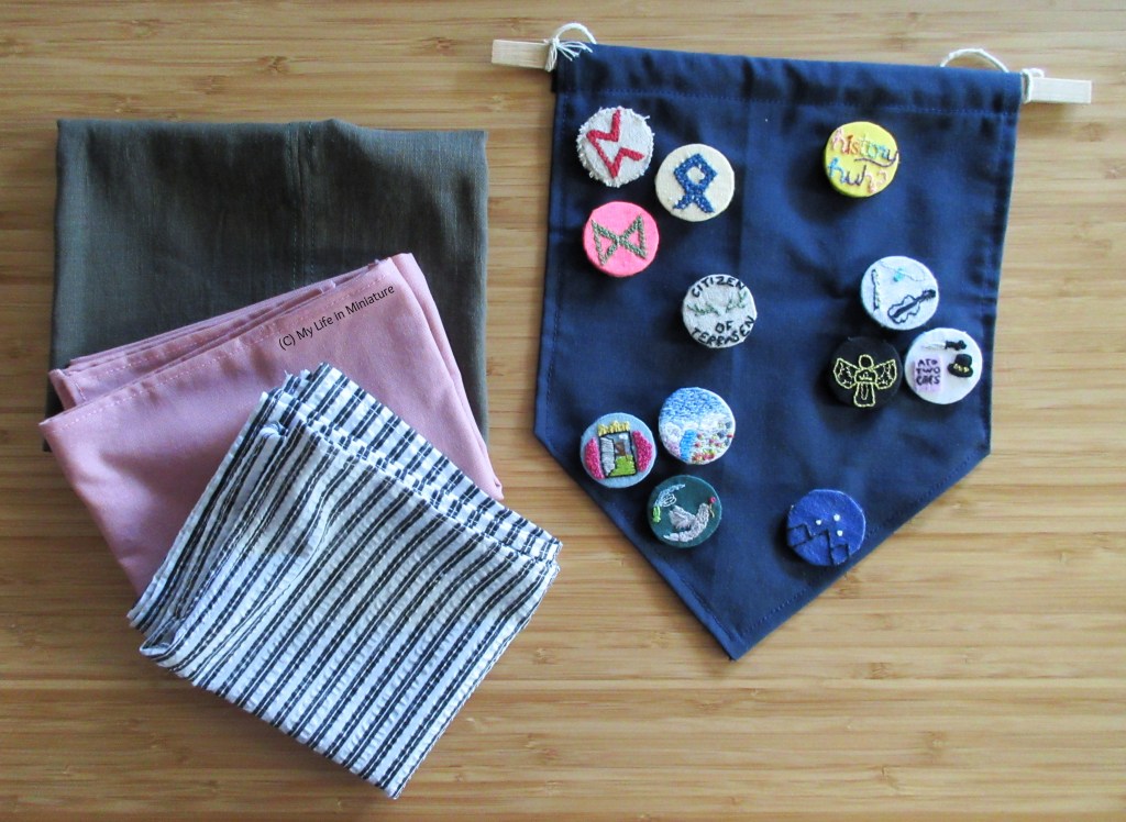 On the left are three folded squares of fabric, slightly overlapping each other. The top one is green, the middle one is pale pink, and the bottom one is black-and-white striped. On the right is a small navy blue banner, mostly covered in embroidered badges. The squares and the banner are on a wood background.