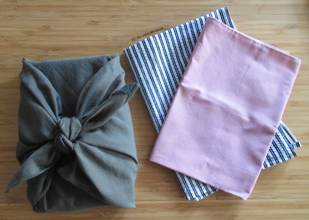 On the wood background, the green furoshiki wrap is tied around a rectangular object. Beside it, the other two wraps are crookedly stacked on top of each other.