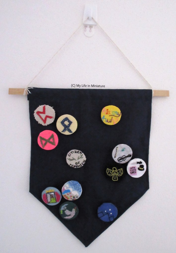 A small navy blue fabric banner, mostly covered in small embroidered badges, hangs from a white hook on a white wall. 
