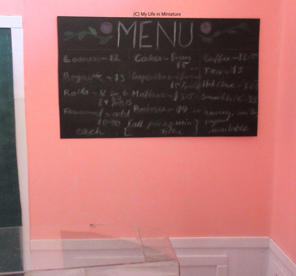 (slightly blurry, shh) image of the menu on the pink wall behind the counter and next to the green-curtained door. The word 'menu' is n all caps across the top, decorated with pink roses and green vines. The prices for various breads, sweet treats, and drinks is listed below in three columns.