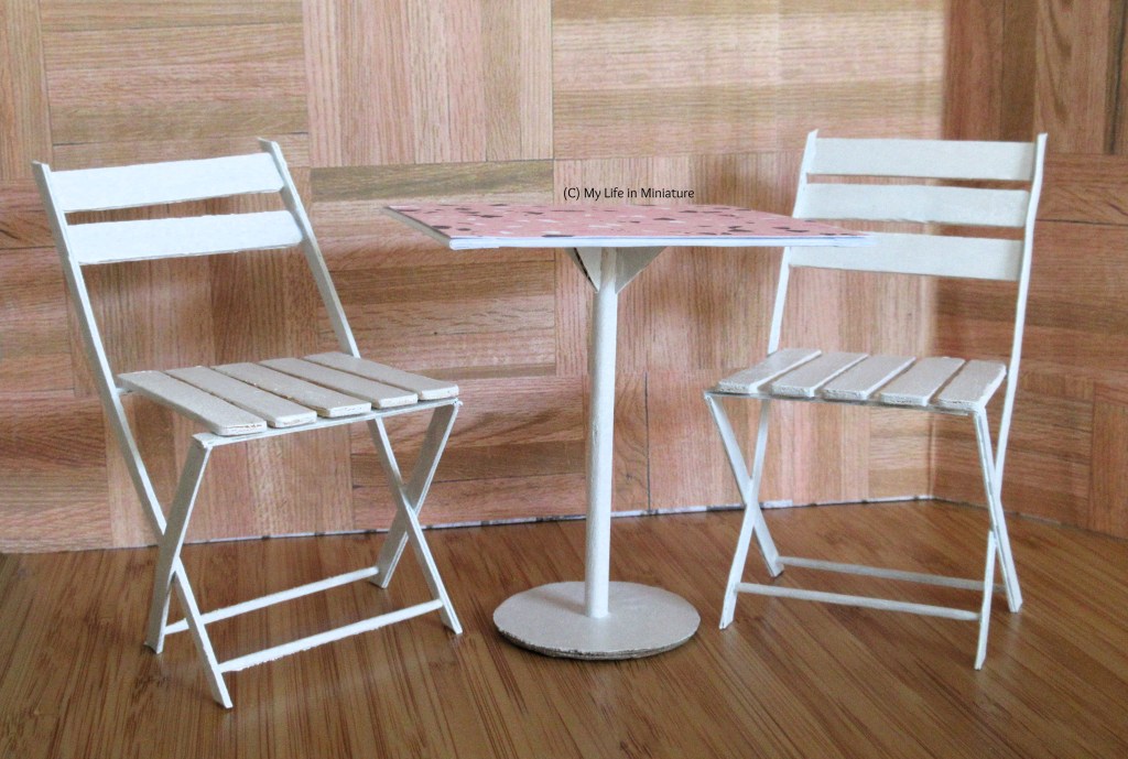 Two white chairs are tucked around a white square table, against a wood backdrop. The chairs look to be foldable café-style chairs, and the table is supported on a central circular dowel and a circular base. The table also has a pink top.