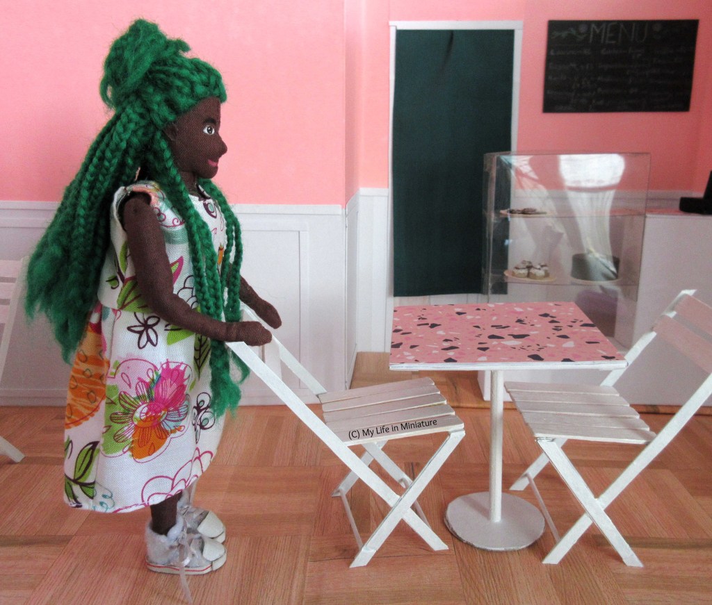 Hazel is in Fierro's, tucking a white chair under a nearby table. A second chair is tucked under the table on the opposite side. Behind the table and chairs is the counter and curtained doorway. Hazel wears a white floral dress with white shoes.