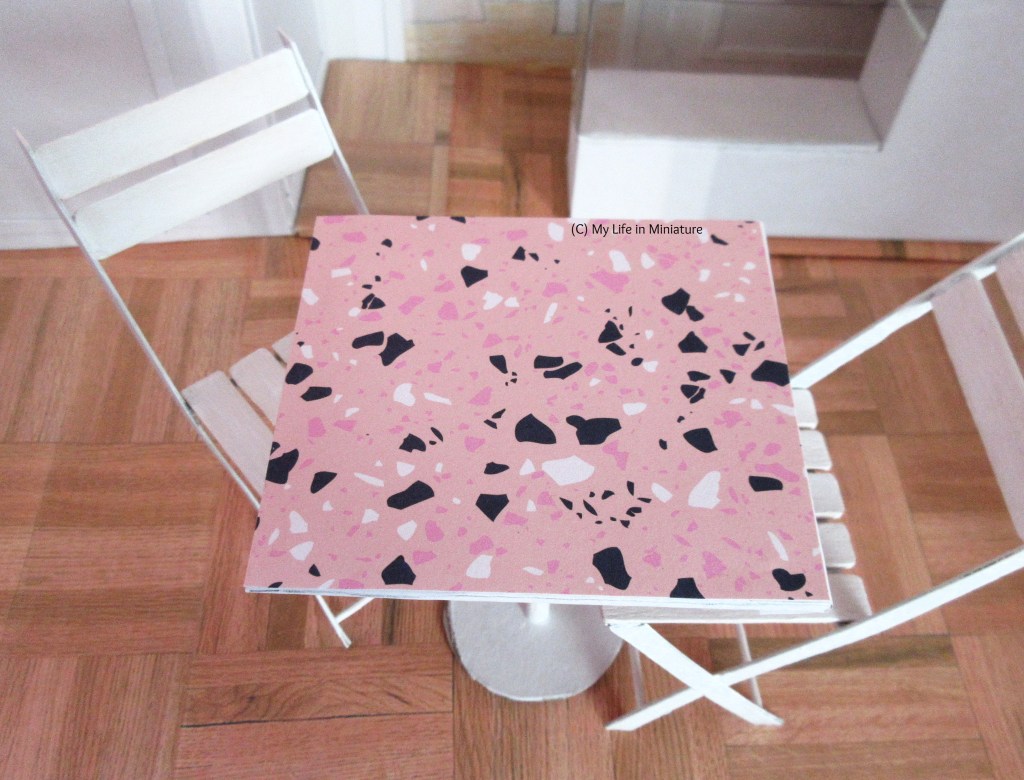 Close-up of the pink top of one of the café tables. It is pale pink, with terrazzo-esque shapes in black, white, and a darker pink scattered randomly. The two white chairs can also be seen, tucked under the table.