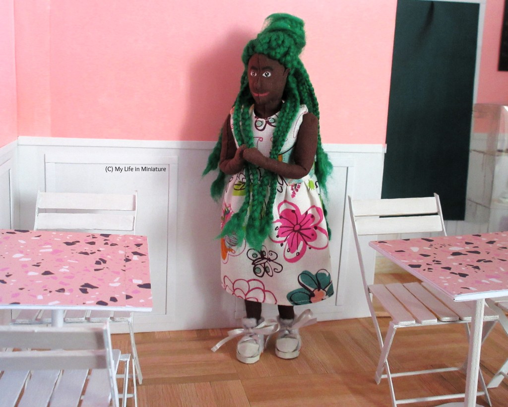 Hazel stands in Fierro's, looking around at the tables and chairs. One set of tables and chairs is against the wall on the left of the image, while the other is to the right of the image. Hazel stands between them, clasping her hands excitedly. She wears a white floral dress with white shoes. 