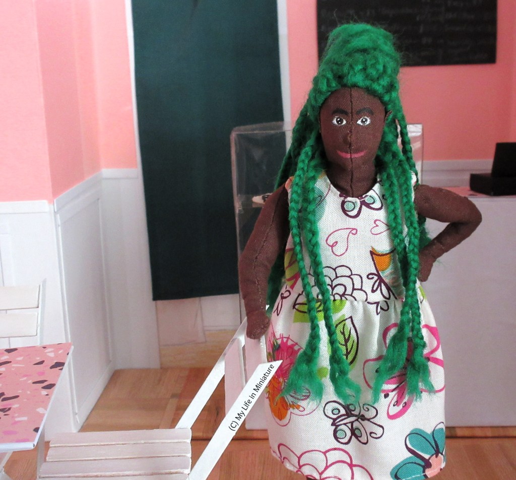 Hazel stands in Fierro's, one hand on the back of a chair and the other on her hip. She smiles at the camera, wearing a white floral dress with some braids pulled forward over her shoulders. Behind her is the front counter and a green-curtained doorway.