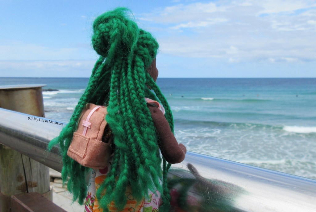 Hazel leans against the railing on the boardwalk, looking away from the camera towards the sea and sky. The backpack is on her back, visible through her parted braids.