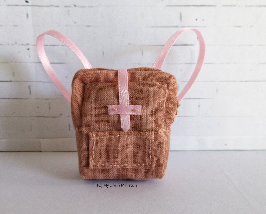A small peach-coloured bag sits against a white brick background. The bag is visually similar to a Fjallraven Kanken bag, however it has pink straps and a pink opening on the front. There is also a small pocket on the front.