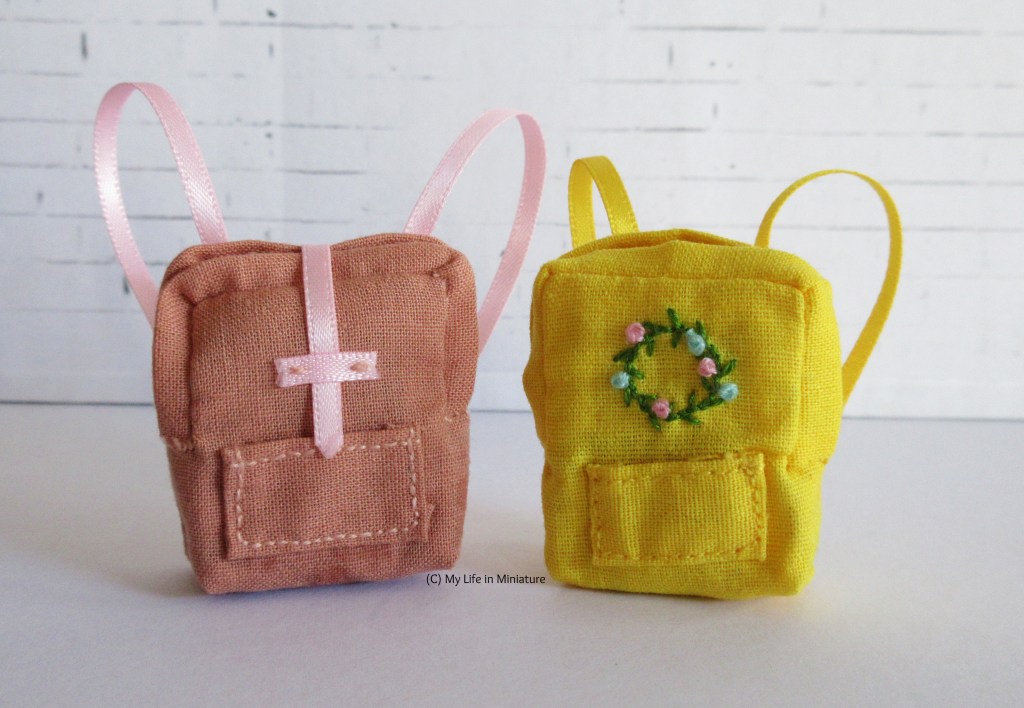 The small peach bag sits next to a very similar yellow bag against a white brick background. The yellow bag has shorter straps and an embroidered floral wreath on the front instead of a closure.
