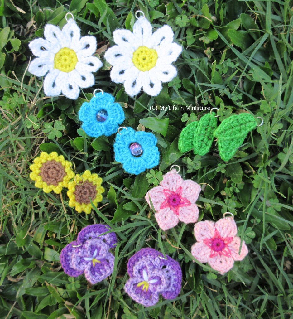 Six crochet earrings sit on grass. There are daisies, green leaves, blue flowers, sunflowers, cherry blossoms, and pansies, all in pairs.