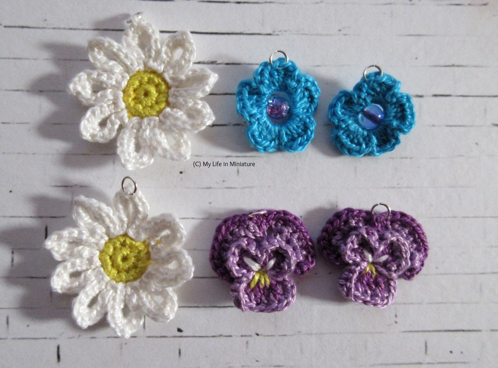 Three pairs of daisy, pansy, and blue flower earrings sit on a white brick background. Each has a small jump ring attached to the top.