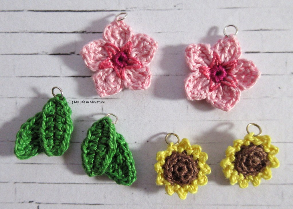 Three pairs of cherry blossom, green leaves, and sunflower earrings sit on a white brick background. Each has a jump ring attached to the top.