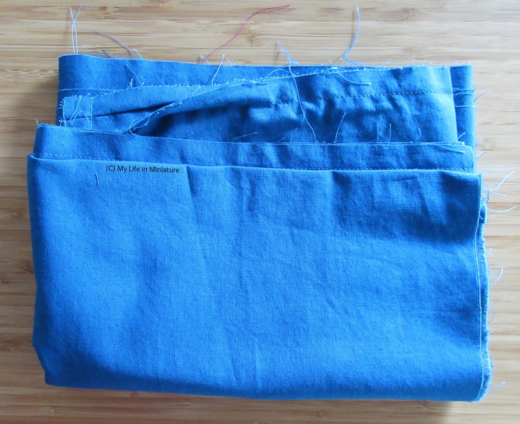 A pair of unfinished sea-blue-green pants is folded and sitting on a wooden background. The way it's folded means it looks more like a large rectangle of fabric, folded up small, at the moment. 
