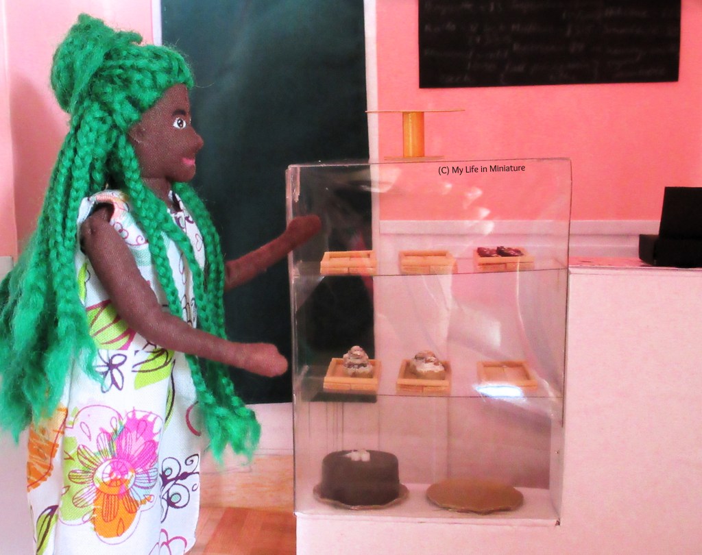 Hazel stands beside the display case/counter, making a 'ta-da!' motion to it. The display case has a gold cake-stand on top, six wooden trays on the next two shelves down, and two gold plates on the bottom shelf. On the trays are biscuits and cinnamon rolls, and one of the gold plates has a chocolate cake on.