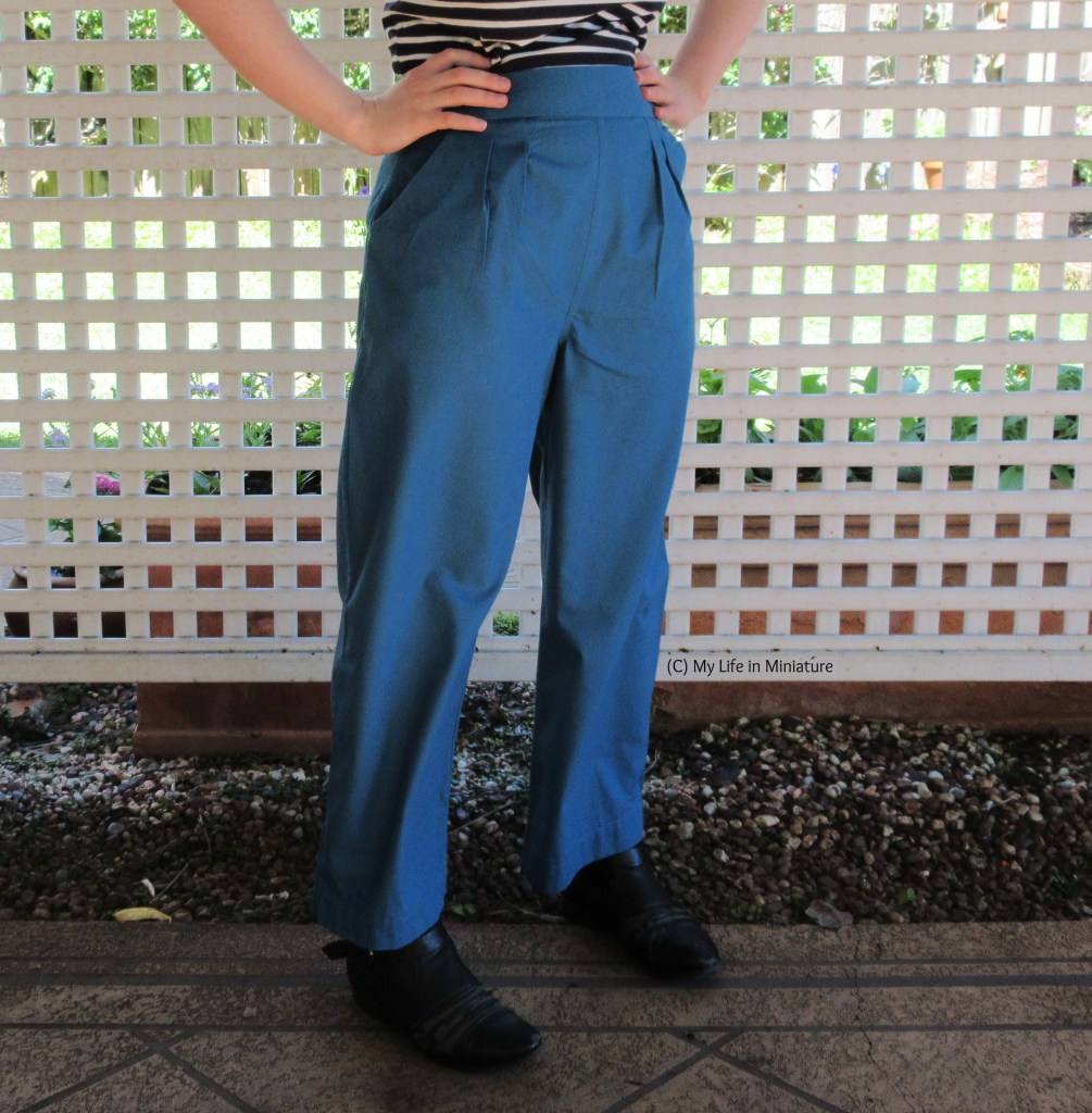 The author stands in front of a white lattice, hands on hips. She wears a pair of sea-blue-green pants with a black-and-white Breton-striped top and black boots. 