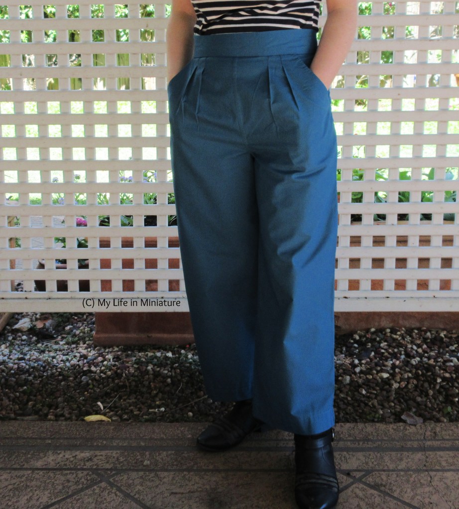 Full=length view of the sea-blue-green pants again. The author stands in front of a white lattice, hands in the two front pockets. The pockets have diagonal openings, and there are four pleats on the front of the pants, under the waistband.