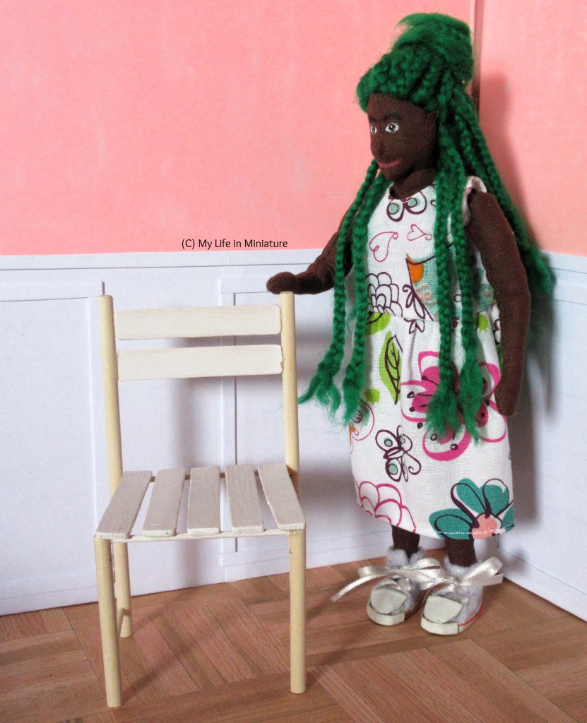 Hazel stands inside Fierro's Bakery, next to an unfinished chair. The chair looks like a regular dining chair, with two slats across the back and five for the seat. The posts are unpainted wood, while the slats are painted white. Hazel wears a white floral dress with white shoes, and has a hand on the back of the chair.
