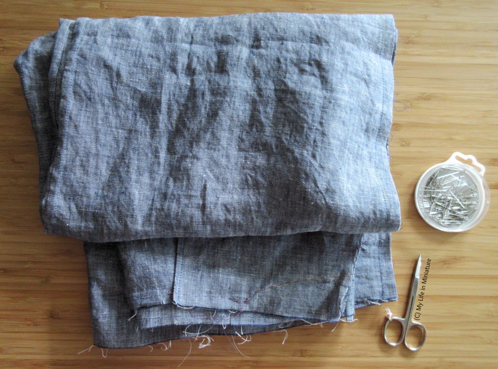 A large, folded-up piece of grey-blue linen sits on a wooden surface beside a pin tin and a pair of embroidery scissors, viewed from above. A few seams are visible, finished and unfinished, as well as a dart.