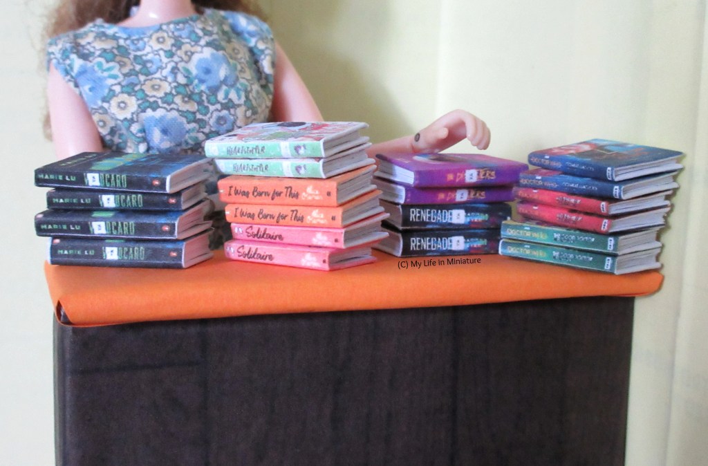 Four stacks of novels sit on the orange-and-wooden front counter in the Palace Library. Sarah is behind the counter in a floral playsuit, about to grab one of the stacks.