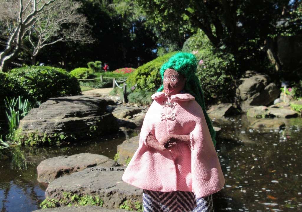 Hazel stands outside, arms clasped in front of her, face turned towards the sun. Behind her are stepping stones across a small river, with the opposite bank in the distance. She wears a pink knee-length cloak with arm slits and a collar over a navy woolen skirt. 