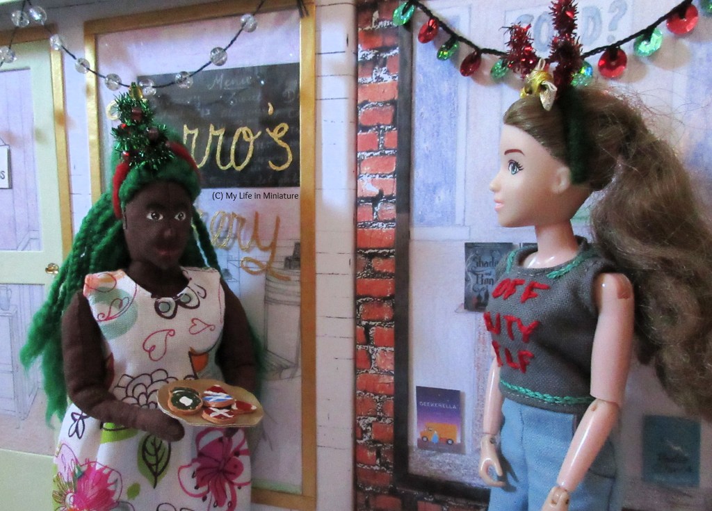 Sarah and Hazel stand outside the Palace Library and Fierro's Bakery, respectively. Sarah looks to Hazel, who has a gold plate of Christmassy biscuits in her hands. She wears a white floral dress and has a headband with a sparkly Christmas tree on. 
