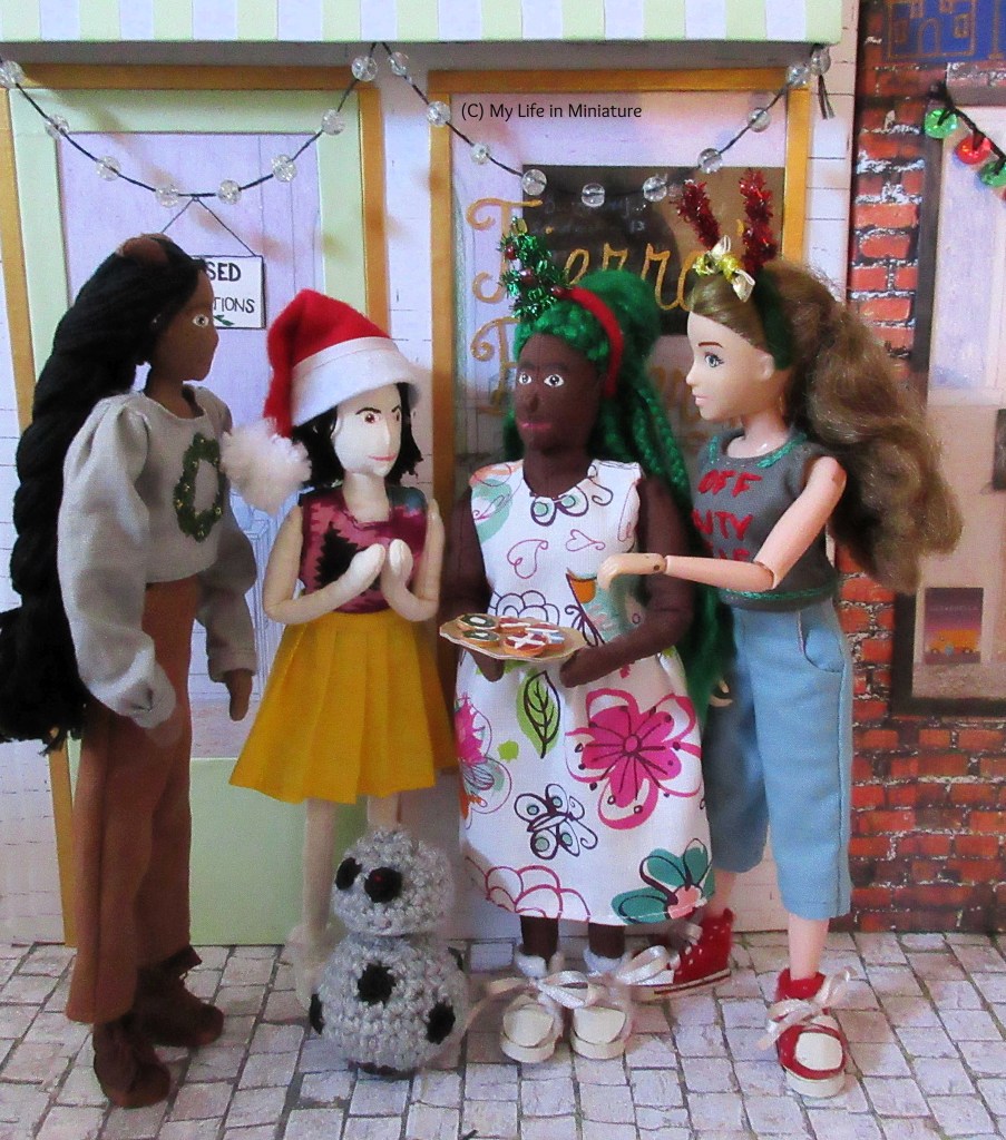 Sarah, Hazel, Tiffany, and Petra stand outside Fierro's Bakery, gathered around Hazel and her plate of Christmas biscuits. Fierro's has a string of 'lights' hanging from the awning. Hazel & Petra look at each other, Hazel answering Petra's question. Tiffany (between Hazel and Petra) looks to Hazel, hands clasped in front of her chest. Sarah reaches around Hazel's shoulder, hand stretched towards the plate. Chip the android is at their feet, face tilted up towards the action. 