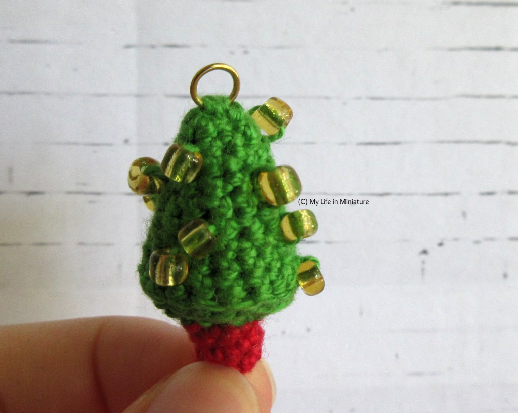 Close-up of one of the Christmas trees. It is a mid-green shade, with a small red trunk at the bottom and a gold jump ring at the top. Gold beads are crocheted into the main tree to look like decorations. 