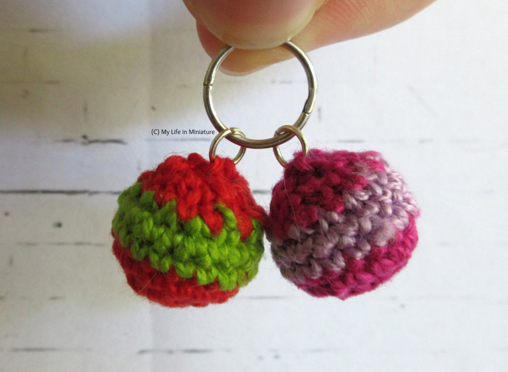 Close-up of the two bauble earrings. Both are striped horizontally in two colours with a gold jump ring at the top. One is red and green, the other is hot pink and pale purple. Both are hanging from one sleeper earring, which the author holds from the top of the image.