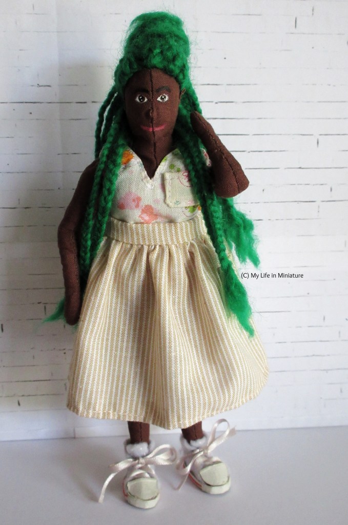 Hazel stands in front of a white brick background. She wears a beige-and-white pinstriped skirt, which is gathered at the waistband and is about midi-length. It is worn with a white floral V-necked top and white shoes. Hazel has a hand pulling two of her braids over her shoulder, and is smiling at the camera.