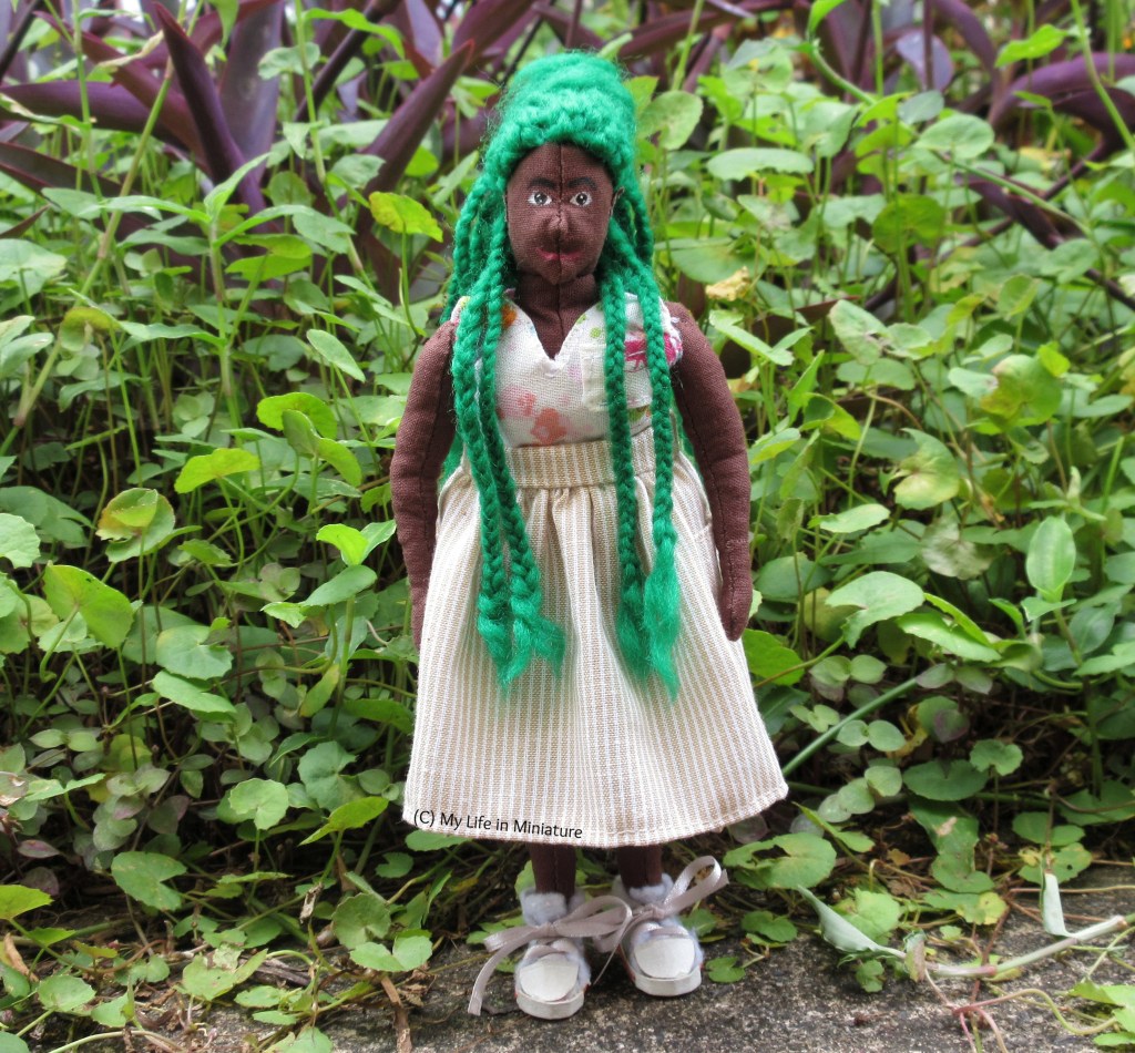 Hazel stands outside, on a concrete path in front of greenery. She wears a beige-and-white pinstriped skirt with a white floral top and white shoes. She has two green braids over her shoulders on each side, her hands at her sides, and is smiling at the camera.