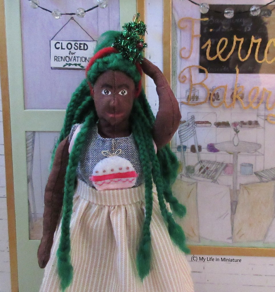Hazel stands outside Fierro's Bakery. She wears a grey-blue top with a bauble on, with a beige-and-white pinstriped skirt, pink backpack, and red headband with a small Christmas tree on. She is reaching up to touch the headband with one hand, and smiling at the camera.