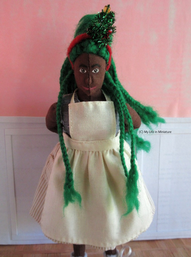 Hazel stands inside Fierro's Bakery, with the half-white-half-pink walls behind her. She faces the camera, tying a cream-coloured apron behind her back. She is looking down slightly, has three braids over her shoulders, and wears a Christmas tree headband. 