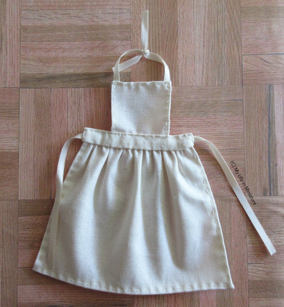 The cream-coloured apron lays flat on a wood background. It has a square to cover the chest and a gathered, midi-length bottom half to protect the legs. There is a strip of cream fabric connecting the gathered bottom half to the flat top half. Straps are attached to the ends of the connecting strip in the middle and the top corners of the upper half. The over-the-neck straps are tied in a knot.
