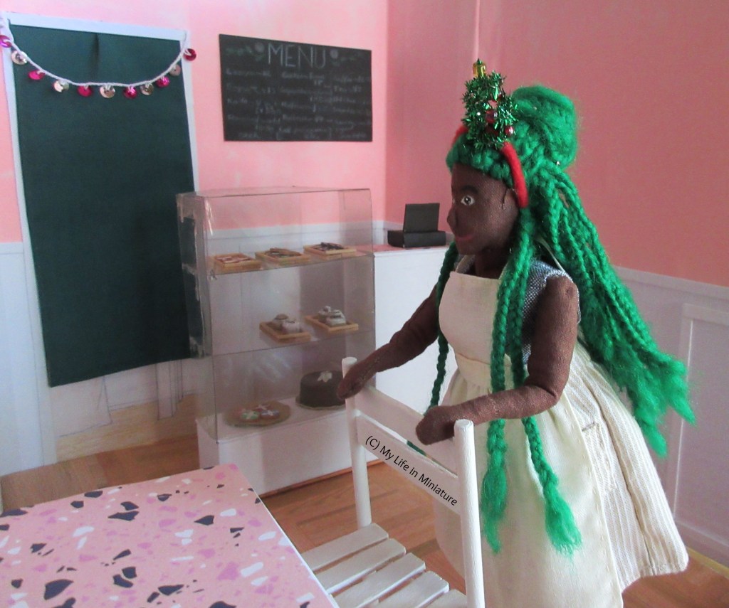 Hazel tucks a chair under a table in Fierro's, wearing the apron over a blue-grey shirt and a cream-and-white skirt, with a Christmas tree headband on her head. Behind her is the counter and the doorway to the back room.