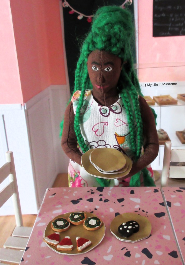 Hazel stands at the two pushed-together tables with four of the six gold plates stacked in her hands. She looks down at the other two plates, which are on the table with biscuits on them. 