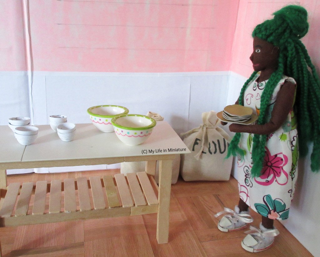 Hazel is standing on the right of Fierro's back room, with a stack of gold plates from out front in her hands. Dominating the room is the work bench, which is covered in 10 bowls of various colours and sizes. To Hazel's right and behind the work bench are two muslin sacks, tied with string.