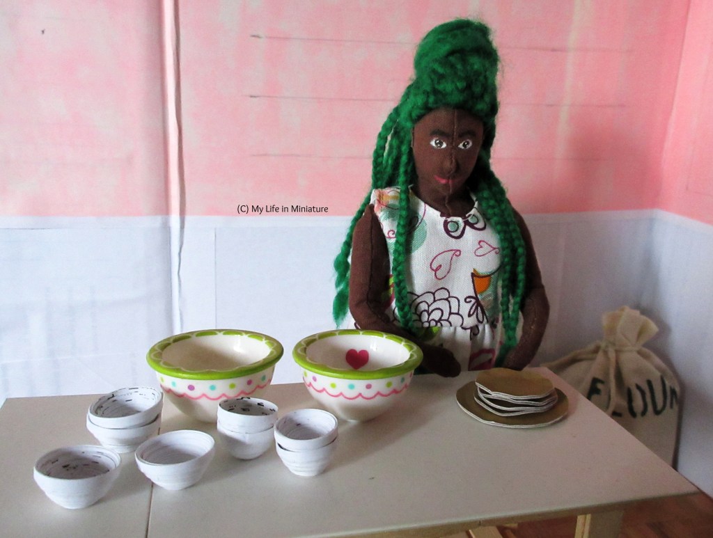 Hazel stands at the work bench in the back room. The gold plates are on the bench in front of her, and she is looking at the bowls that cover the rest of the surface. Just behind her, the flour sack is visible.