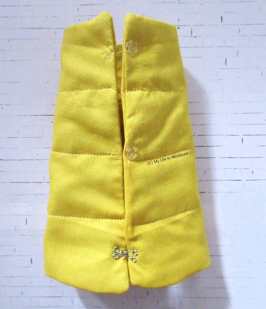 The reverse side of the yellow armband, photographed on a white brick background. A hook and eye closure is sewn near the elbow end of the armband. In the centre and near the wrist of the armband are two sets of clear press studs/snaps.