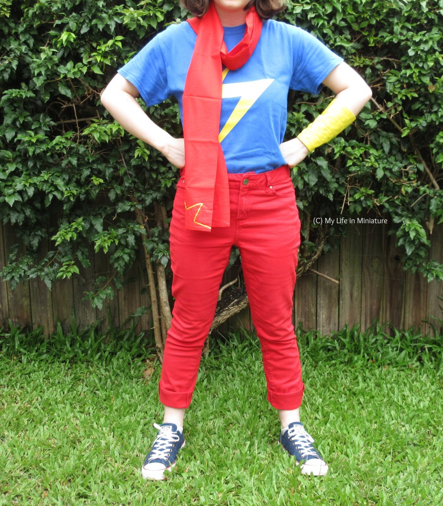 The author stands in front of a hedge on grass. She wears a 'casual cosplay' of the comics character Ms Marvel. This consists of a blue shirt with a yellow zigzag on, red jeans cuffed at the bottom, denim Converse shoes, a red scarf with yellow detail on the end, and a yellow fabric cuff/gauntlet on her left arm. She stands with her hands in fists on her hips, in that classic Wonder Woman pose. 