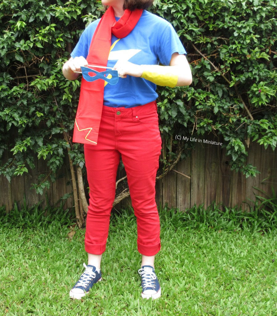The author stands against a hedge on grass, wearing the relaxed Ms Marvel cosplay outfit. She faces the camera, angled slightly to the left, with the mask in both hands. Her pose is alert, and it looks like the mask is about to go on her face (which isn't in shot). 