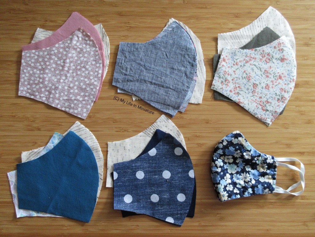 Six masks are laid out on a wood background in two rows of three. Only the bottom right one is actually complete - the other five are just the pieces cut out in various different fabrics. There is a pink one, two in shades of green, and two in shades of blue. Each has three layers. 