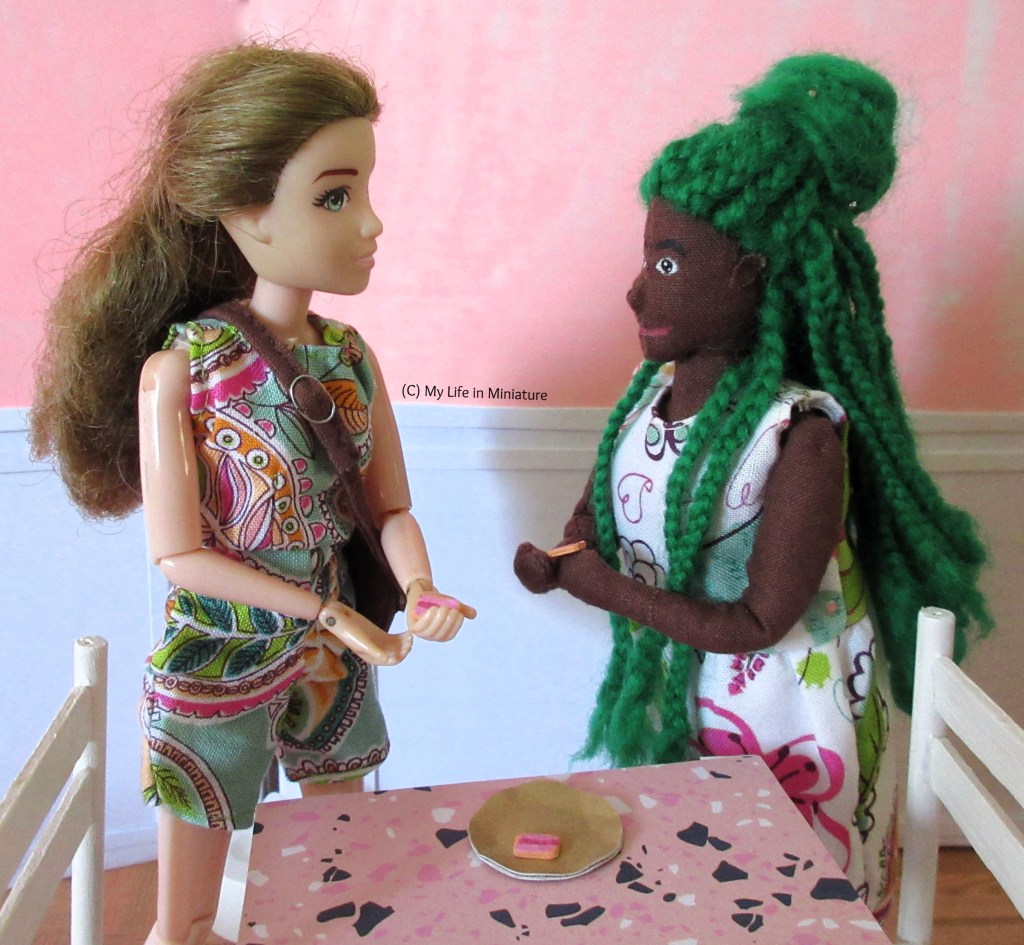 Sarah and Hazel stand beside a Fierro's table, in discussion. Both hold an Iced Vovo with a bite taken out of it. There is one biscuit left on the plate. 