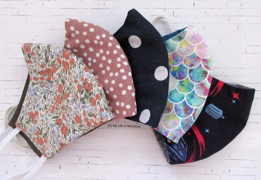 Five masks are fanned out in a white brick background. Left to right, the designs are green and floral, dusky pink with white polka-dots, navy with bigger white polka-dots, pink iridescent scales, and navy Doctor Who fabric. 